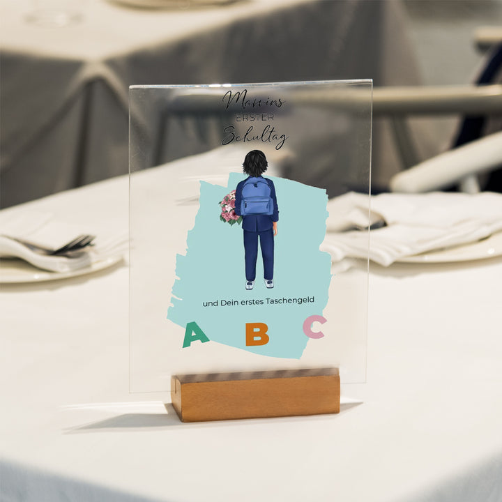 Personalisierte Acryltafel für den ersten Schultag mit Illustration eines Schulkindes, Buchstaben ABC und Holzständer. Individuelles Geschenk zur Einschulung auf einem gedeckten Tisch präsentiert.