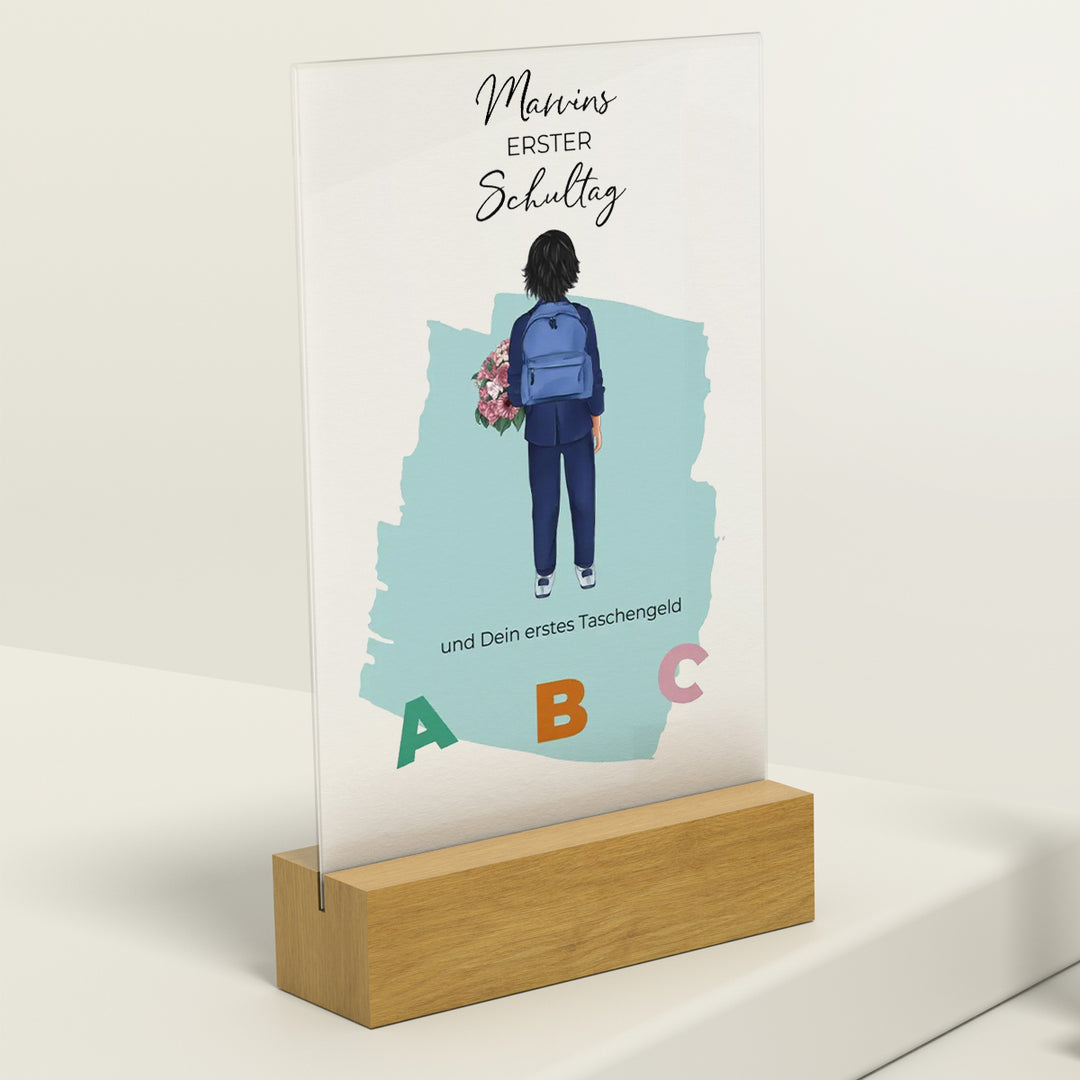 Personalisierte Acryltafel für den ersten Schultag, zeigt Silhouette eines Kindes mit Rucksack und Blumen vor hellblauem Hintergrund, ABC-Buchstaben und Text "Marvins erster Schultag", auf Holzständer präsentiert