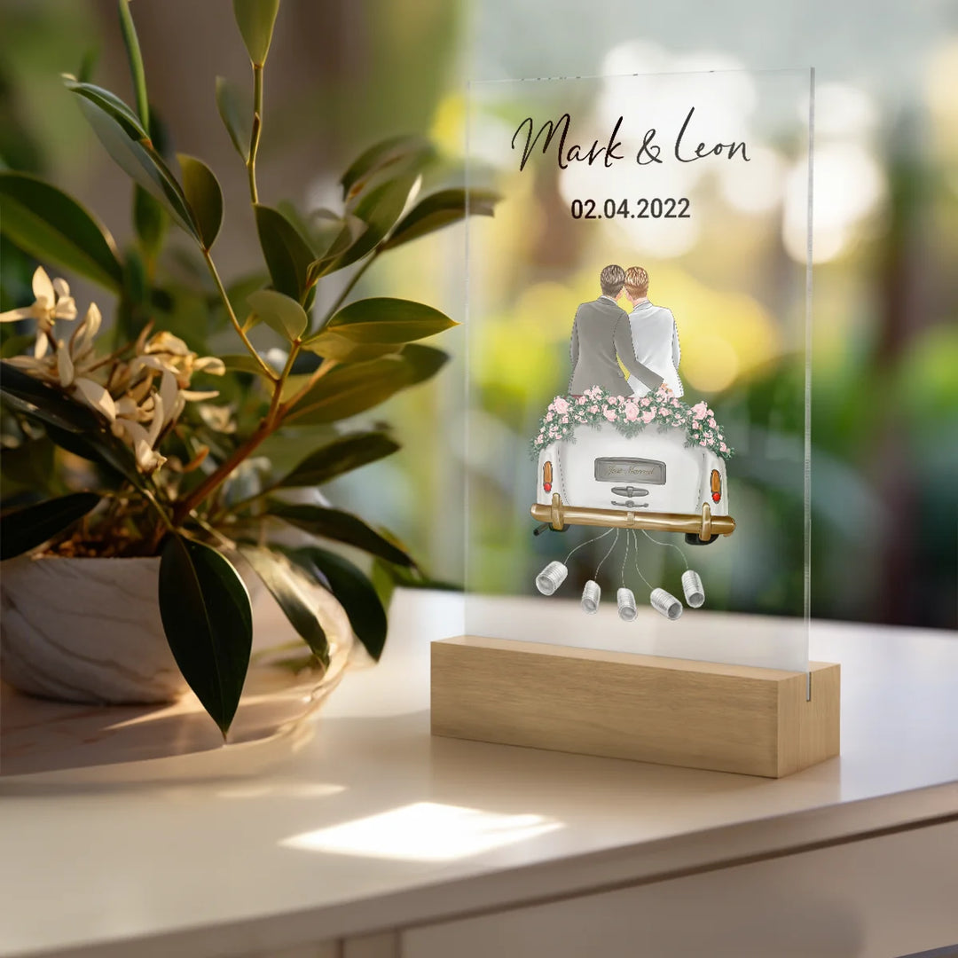 Personalisierte Acryltafel als Geldgeschenk zur Hochzeit mit Namen und Datum, zeigt ein frisch vermähltes Paar in einem geschmückten Auto. Dekorative Zimmerpflanze im Vordergrund. Elegantes Hochzeitsgeschenk für gleichgeschlechtliche Paare.